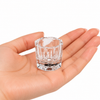 Small clear glass mixing dish for professional lash and brow tinting, held in the palm of a hand on a white background